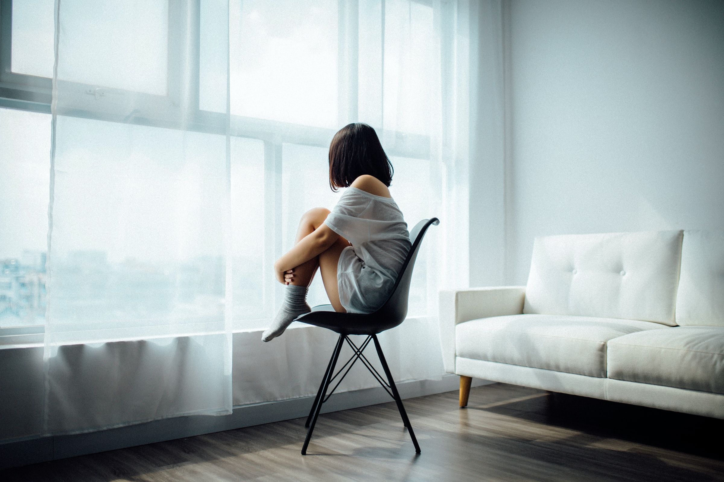 Eine Frau sitzt auf einem Stuhl und hält ihre Beine zusammen während sie aus einem Fenster schaut