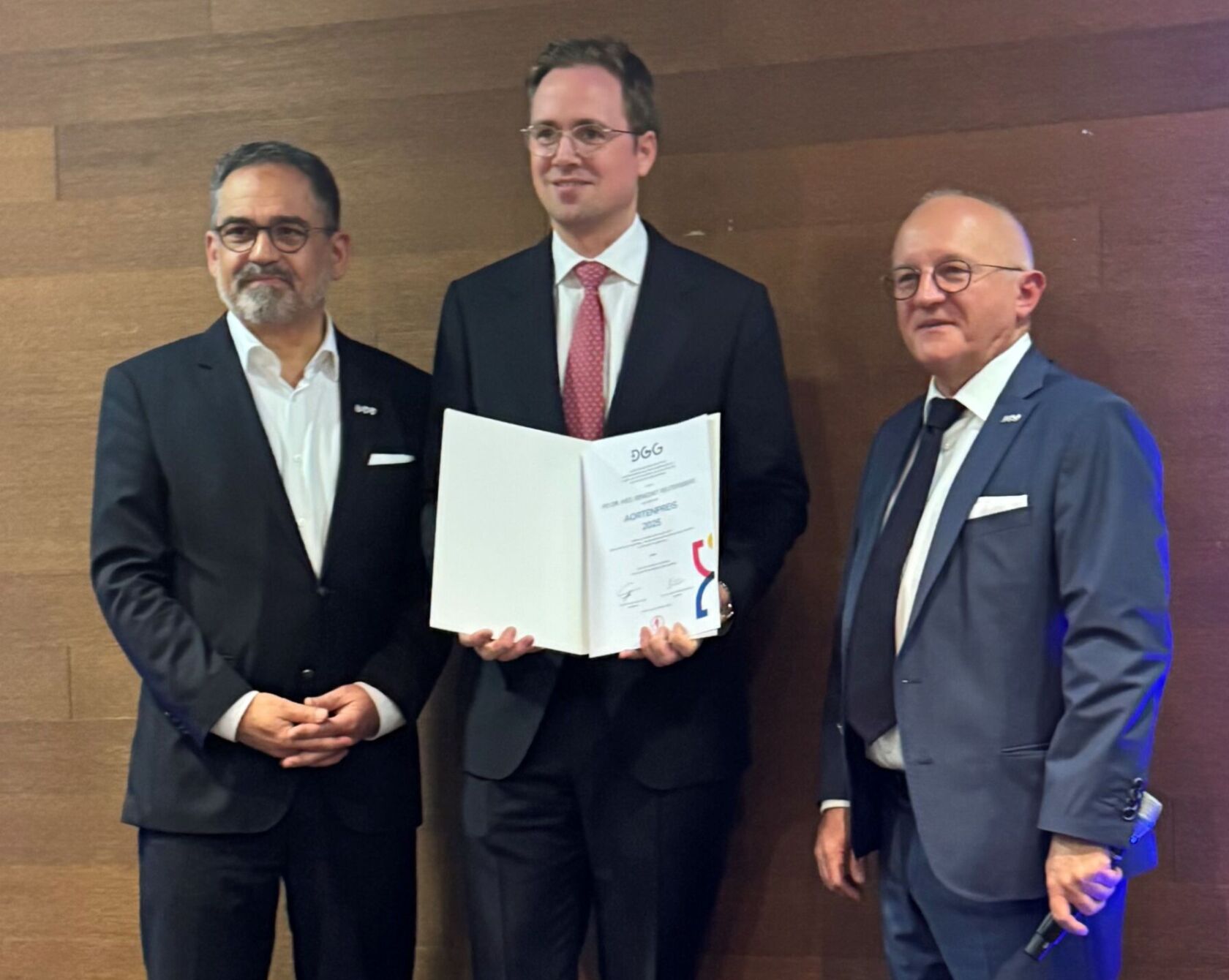 Dr. Reutersberg hält seinen Preis in die Kamera, zwei Personen stehen daneben.