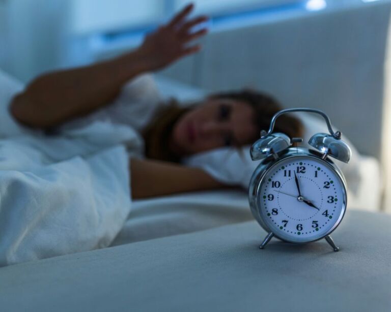 Wecker im Vordergrund, zeigt vier Uhr an, Frau wach im Hintergrund