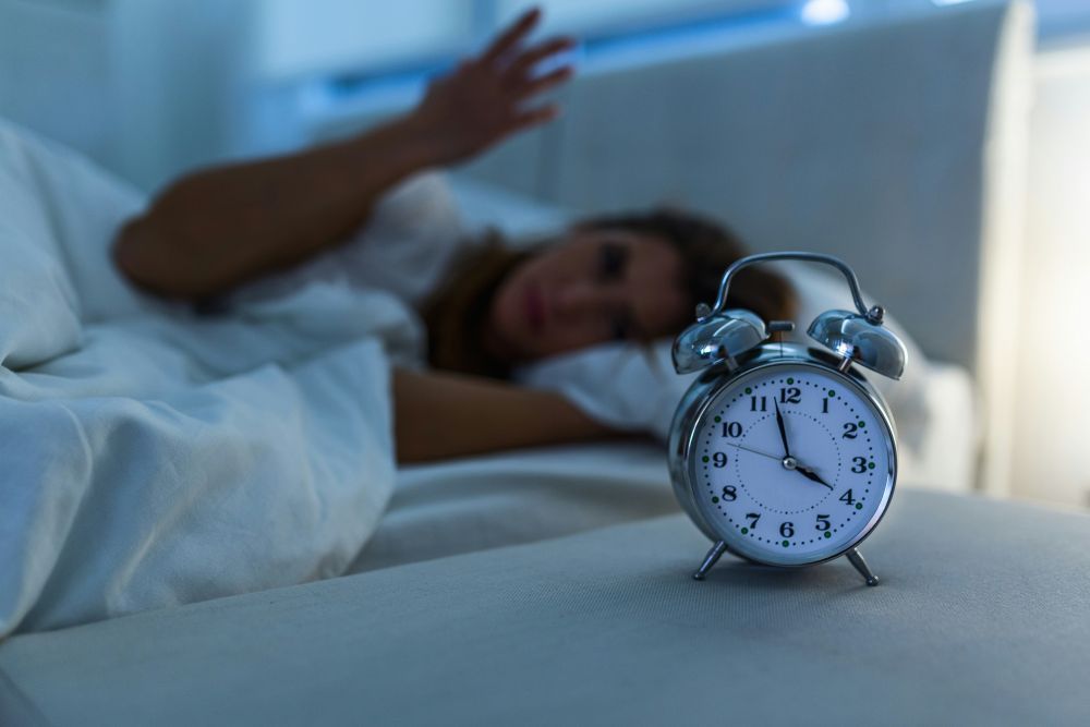 Wecker im Vordergrund, zeigt vier Uhr an, Frau wach im Hintergrund